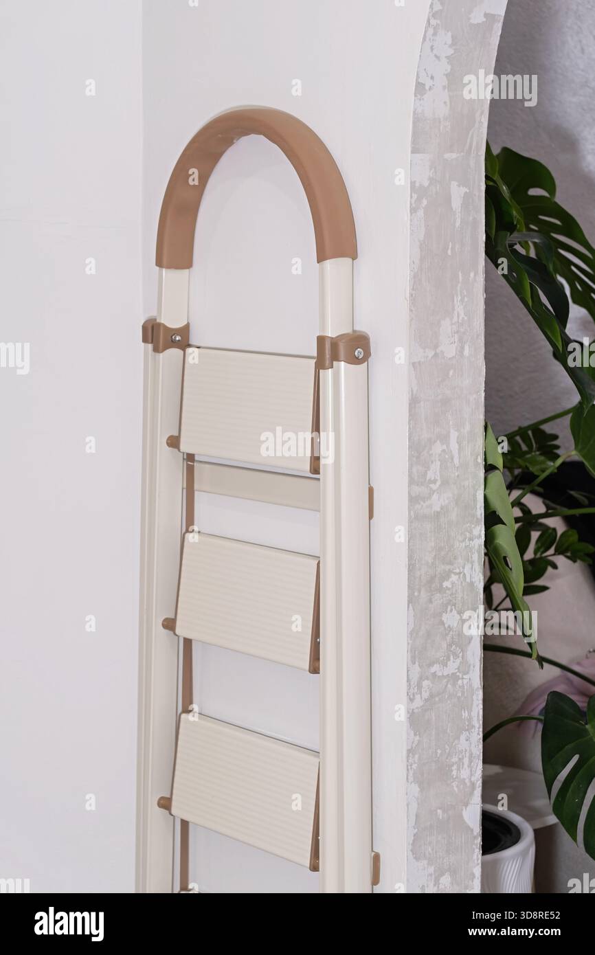 Beige klappbare Trittleiter an weißer Bogenwand mit grüner Zimmerpflanze in gemütlichem, modernem Wohnraum, symbolisiert Renovierung, heimwerkerarbeit und Haushaltsgestaltung Stockfoto