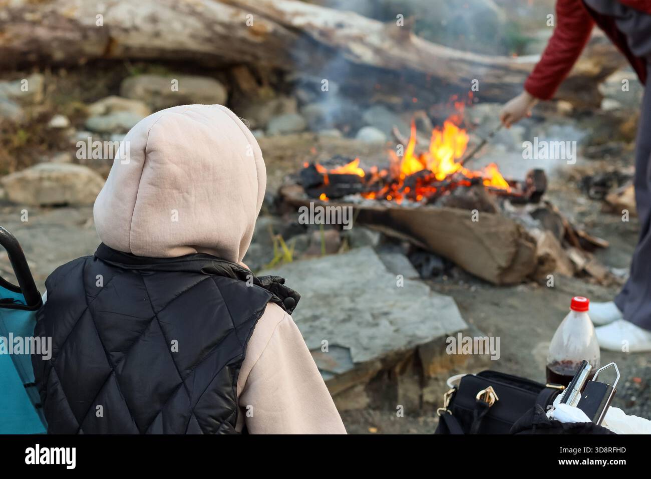 Eine Person in roter Jacke fügt einem Lagerfeuer Holz hinzu, während eine andere in Kapuze und Weste neben einem blauen Campingstuhl an einem felsigen Uferufer sitzt. Stockfoto