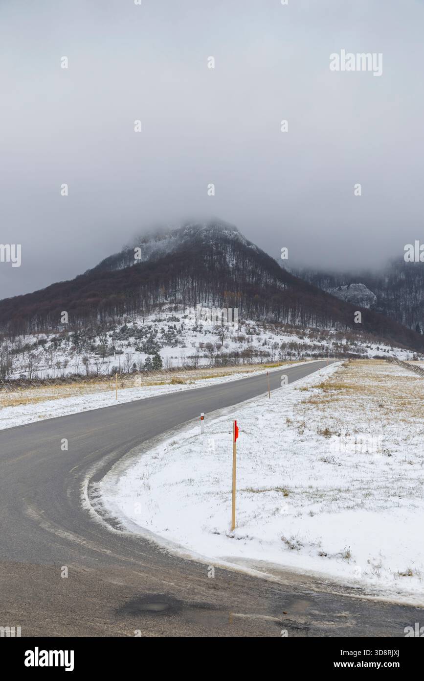 Gekrümmte Asphaltstraße, die zu einem Berg führt, der unter einem bewölkten Himmel mit Schnee und Nebel bedeckt ist Stockfoto
