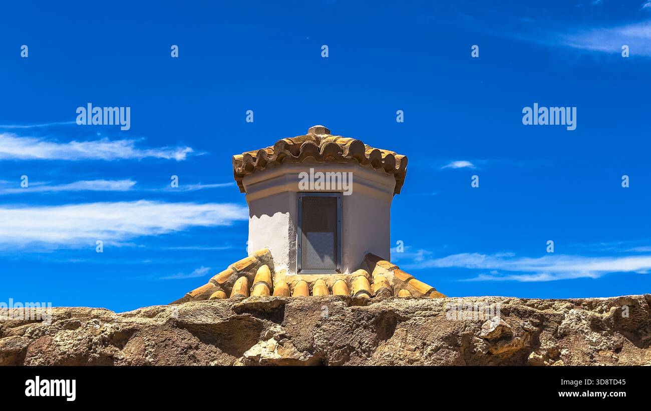 Aus der Nähe des rustikalen Dachs mit verwitterten Tonziegeln und einem kleinen weißen Lüftungsturm vor einem leuchtend blauen Himmel mit weichen Wolken. Stockfoto