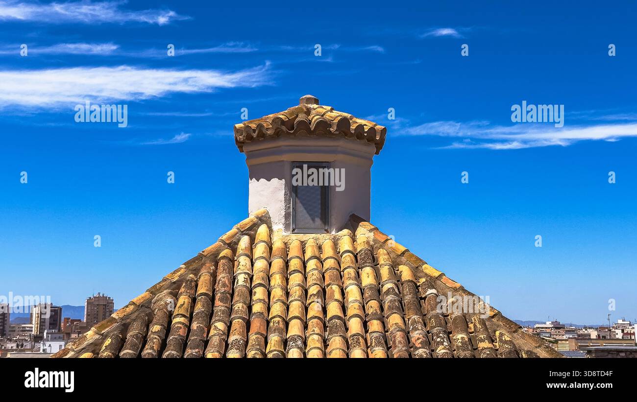 Traditionelles mediterranes Dach mit rustikalen Lehmbliesen und kleinem weißen Schornsteinturm vor hellblauem Himmel und weichen Wolken an sonnigen Tagen. Stockfoto
