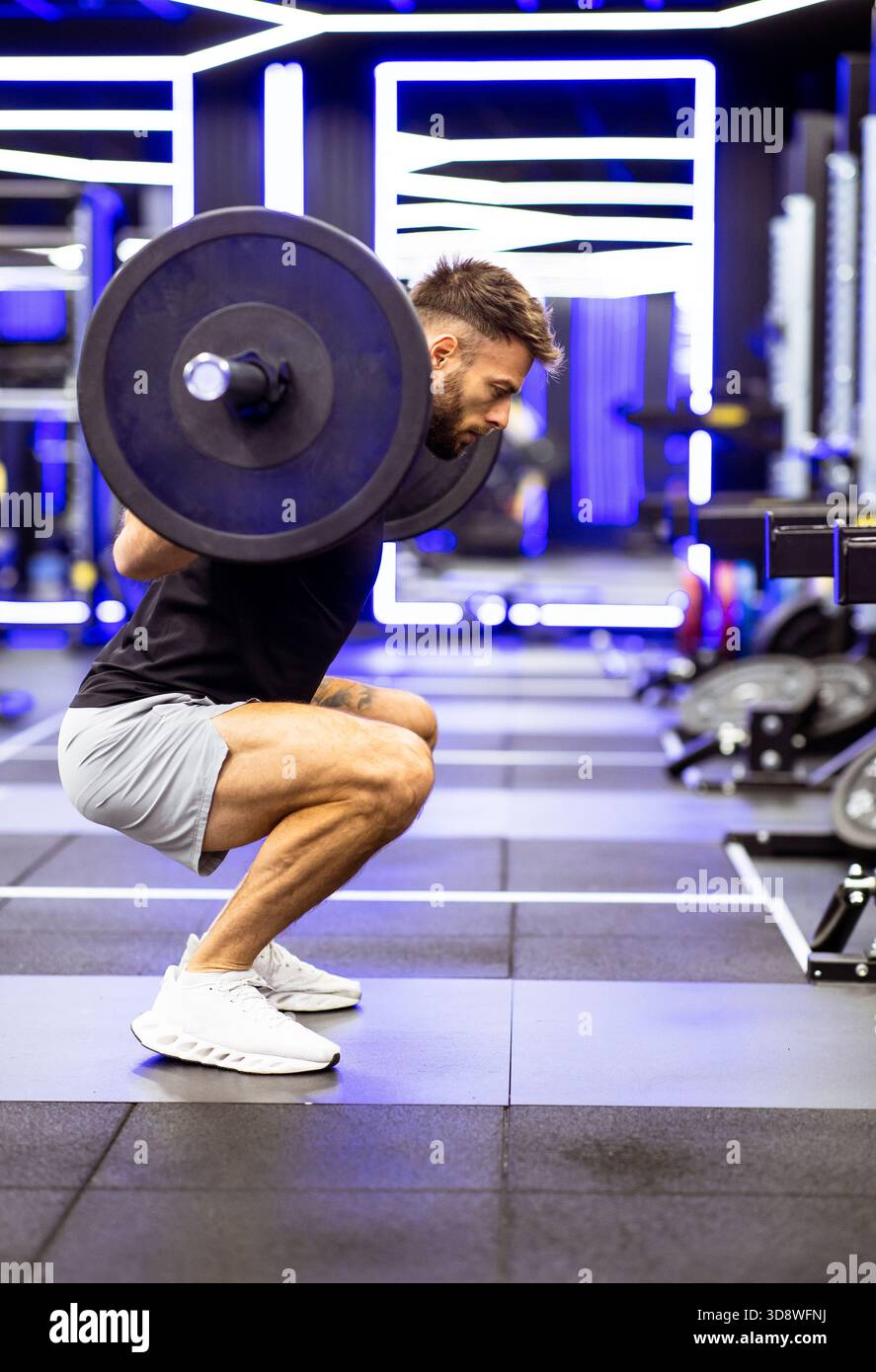 Ein Mann führt in einem eleganten und lebhaften Fitnessstudio eine Hocke mit einer schweren Langhantel aus und zeigt beim Workout Engagement und Kraft. Stockfoto