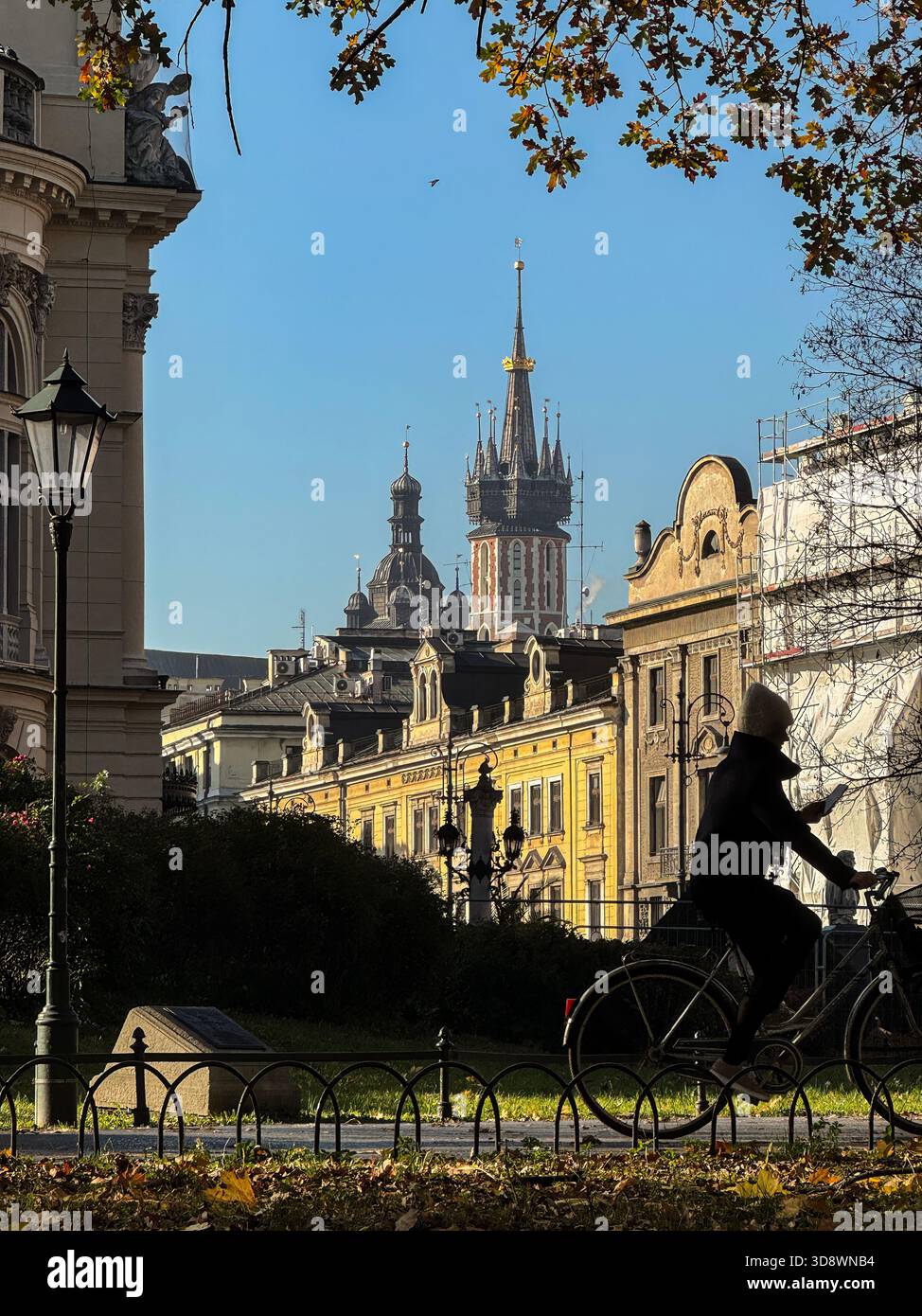 Eine sonnendurchflutete Straße in der Altstadt von Krakau mit den Türmen der historischen Marienkirche im Hintergrund, vorbeifahrenden Radfahrern und warmen gelben Steinhäusern. Klasse Stockfoto