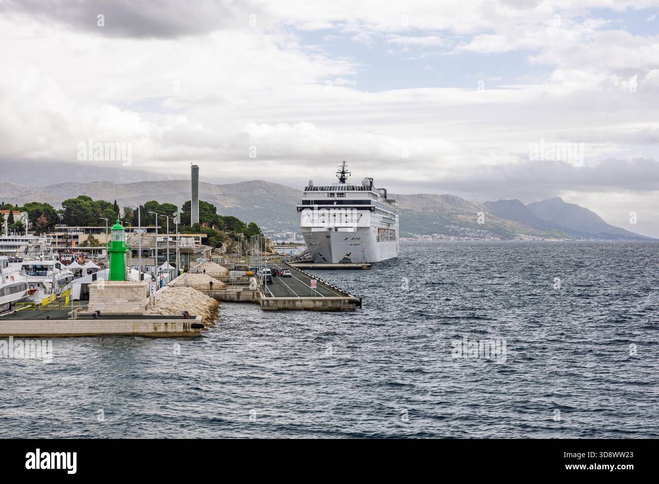 SPLIT, KROATIEN, 10.25.2025: Ein großes weißes Kreuzfahrtschiff liegt an einem belebten Hafen neben einem leuchtend grünen Leuchtturm. Steinsteg, vertäute Boote und entfernte m Stockfoto