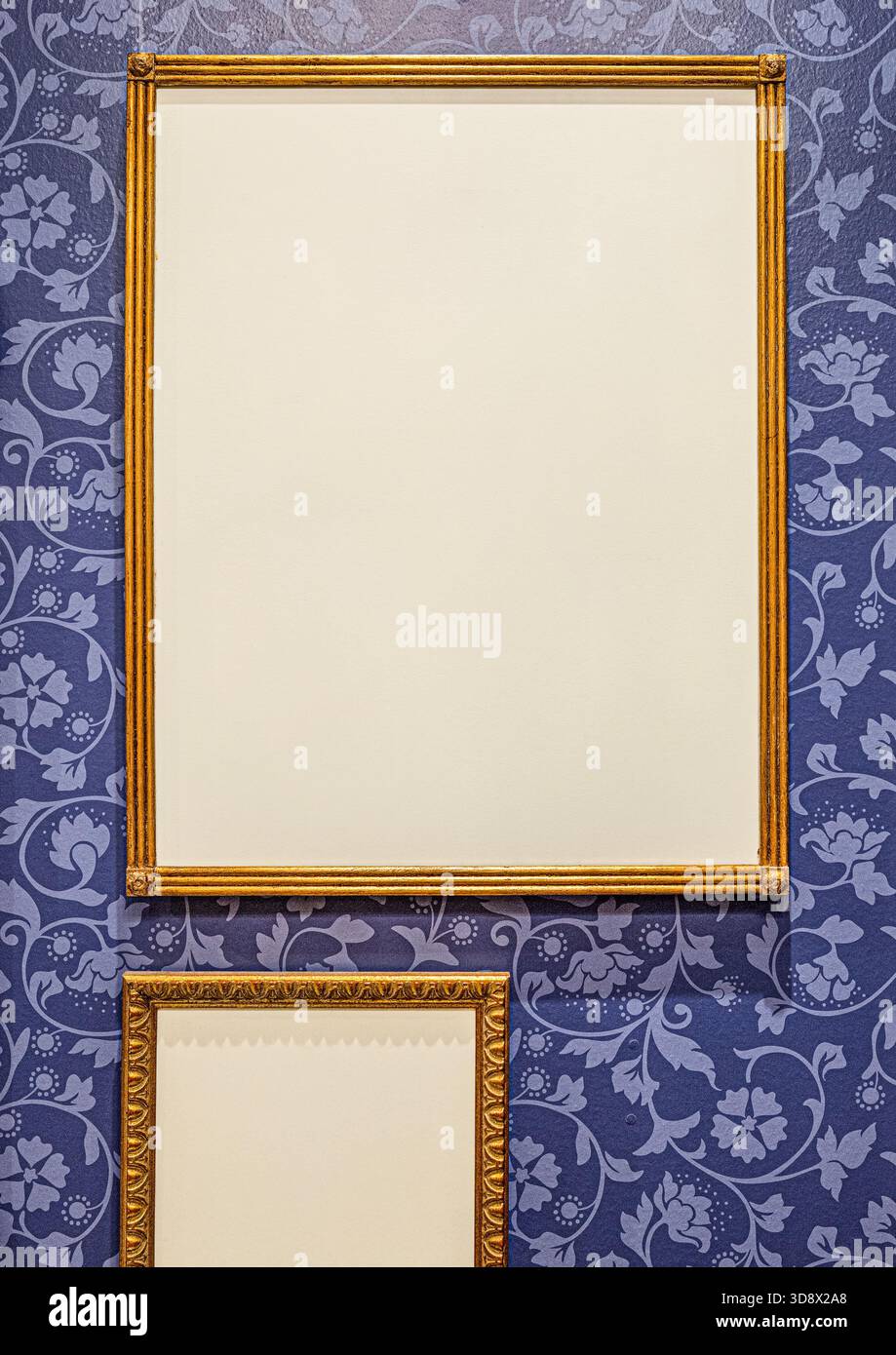 Zwei leere goldene Bilderrahmen hängen an einer dunkelblauen Wand mit Vintage-floraler Tapete. Stockfoto