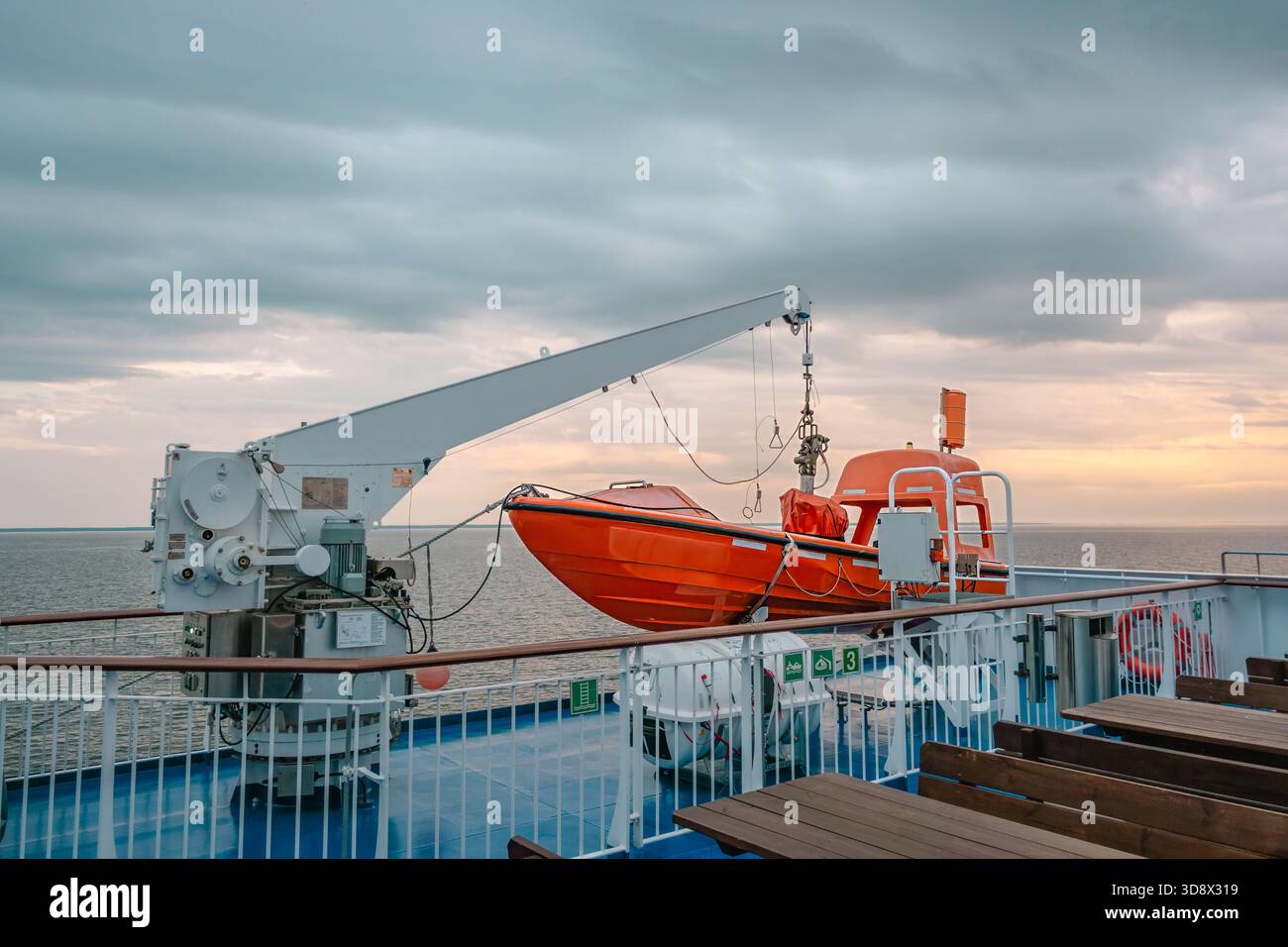 Großes orangenes Rettungsfloß, das von einem weißen Kran auf dem Oberdeck einer Fähre gehoben wird und bei Sonnenuntergang unter dramatischen Wolken über die Ostsee segelt, maritimes saf Stockfoto