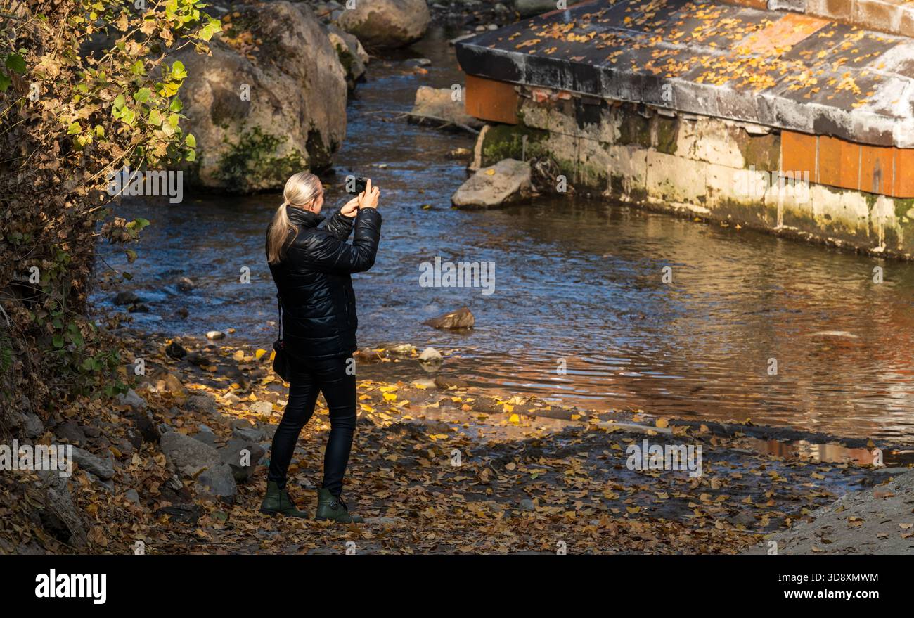 Jerewan Armenien 10.25.2025. Eine Frau fotografiert einen Fluss. Das Wasser ist ruhig und die Blätter auf dem Boden sind braun Stockfoto