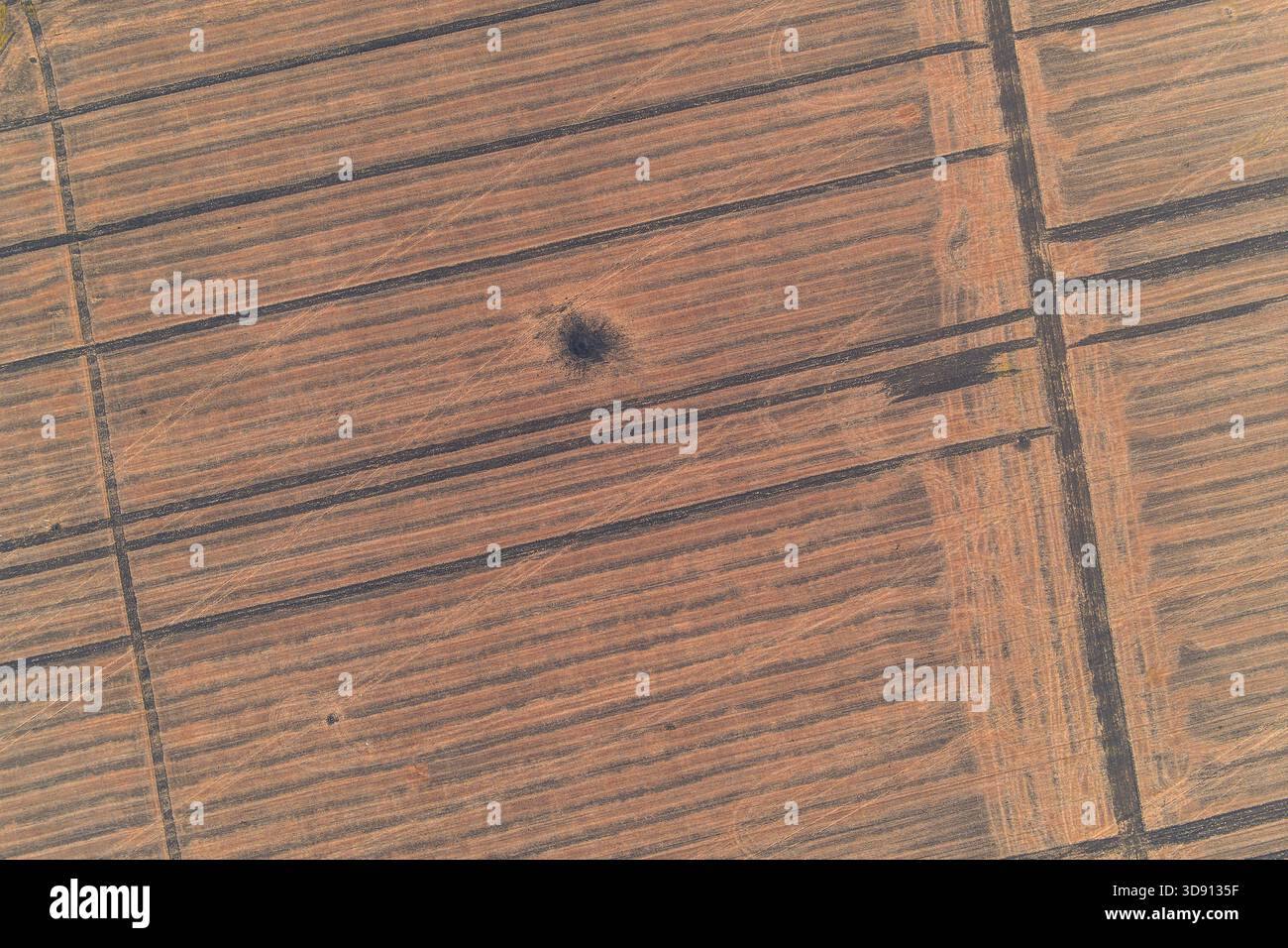 Abstrakter Drohnenschuss, der das einheitliche Muster eines ausgedehnten gepflügten Feldes erfasst, das von einem einzigen dunklen kreisrunden Muschelkrater unterbrochen wird. Die minimalistische Komposition veranschaulicht eindrucksvoll die Narben von Konflikten. Stockfoto