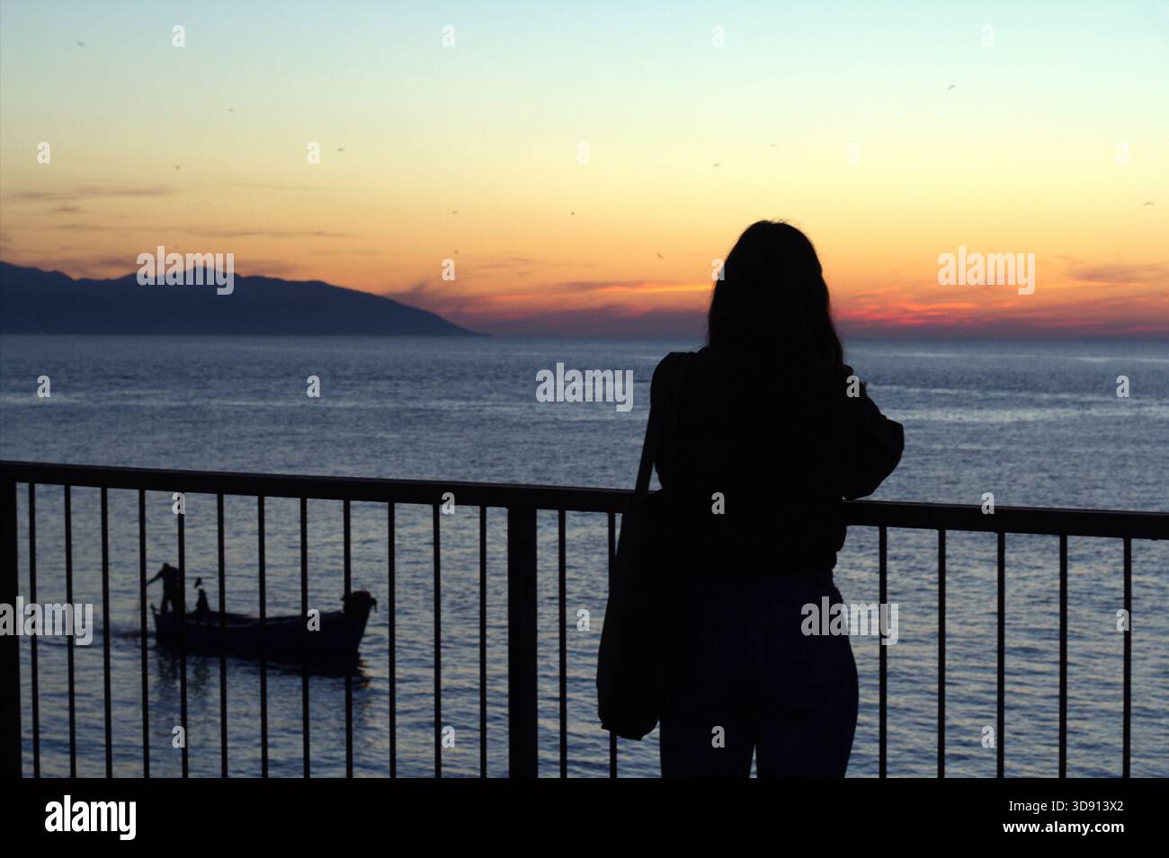 Einsame Silhouette bei Sonnenuntergang über dem Meer Die Silhouette Einer Frau steht auf einem Balkongeländer und blickt auf das ruhige Meer, während die Sonne untergeht Stockfoto