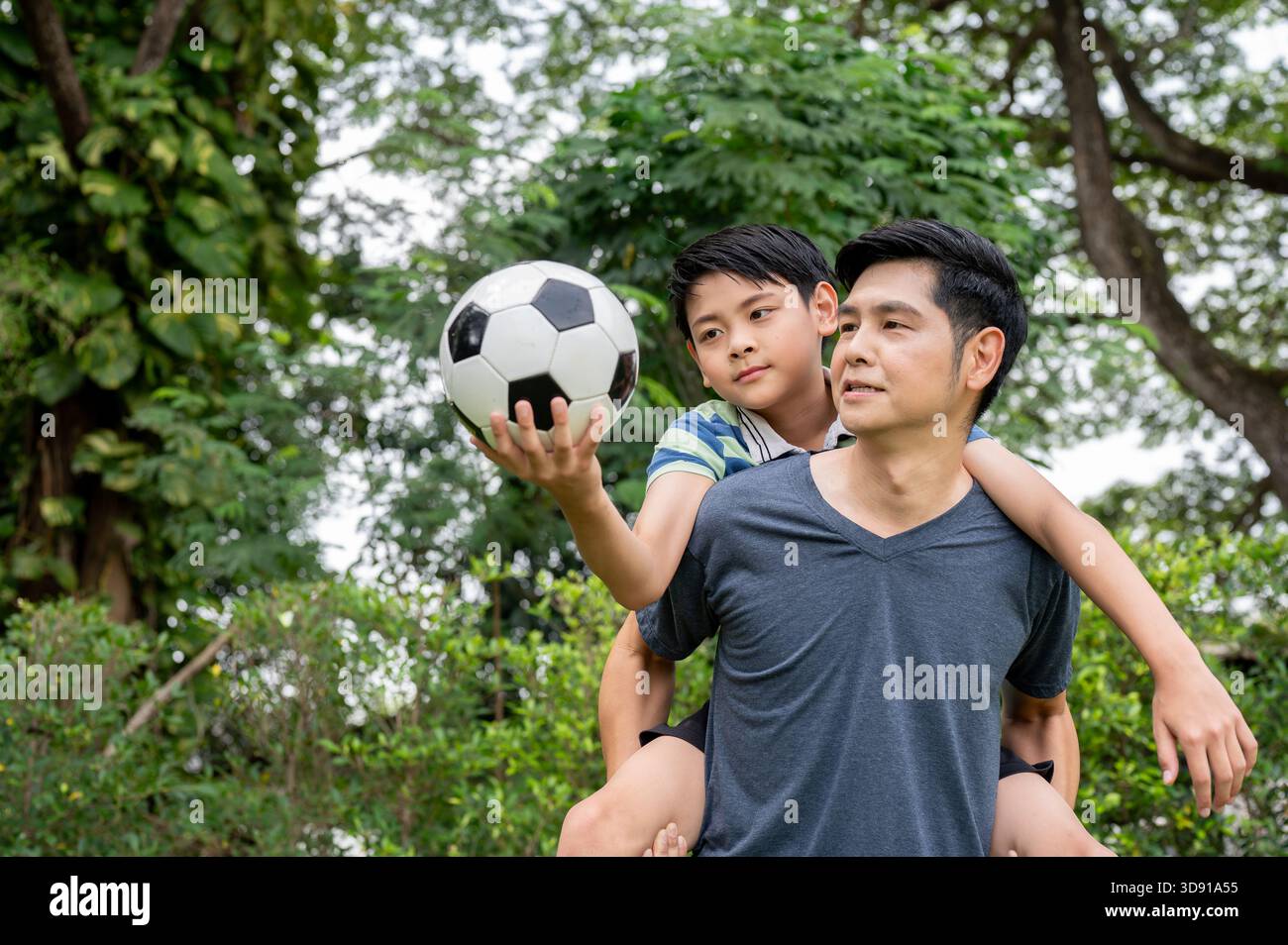 Vater trägt einen Sohn, der einen Ball auf dem Rücken hält, nachdem er zusammen Fußball oder Fußball in ihrem Garten gespielt hat. Familienverbundenheit, Sportaktivitäten. Stockfoto