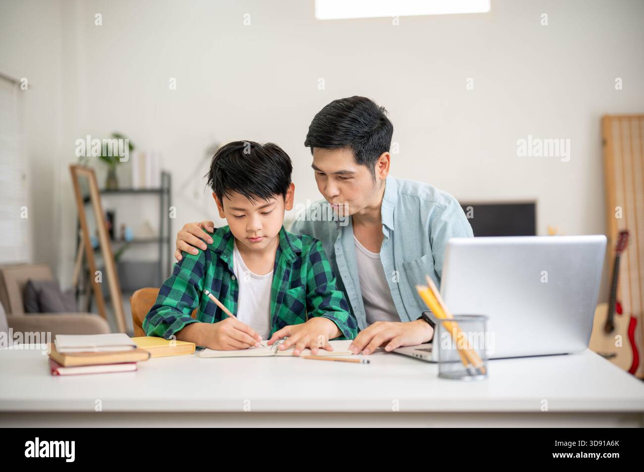 Vater sieht glücklich aus mit dem Arm auf der Schulter seines Sohnes, während sein Sohn Hausaufgaben am Tisch macht. Grundschüler, Kindererziehung, Family Bounding. Stockfoto