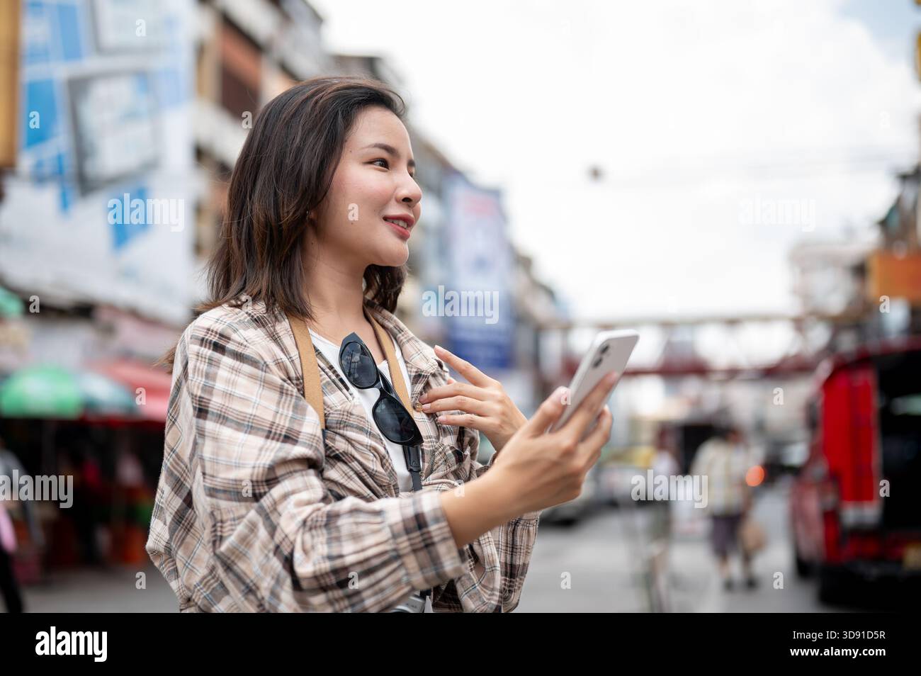 Asiatische Touristenfrau im Scotch-Muster-Hemd, das Telefon hält und auf einem lokalen Markt nach vorne blickt. Fußgängerstraße, Ausflug in die Altstadt. Stockfoto