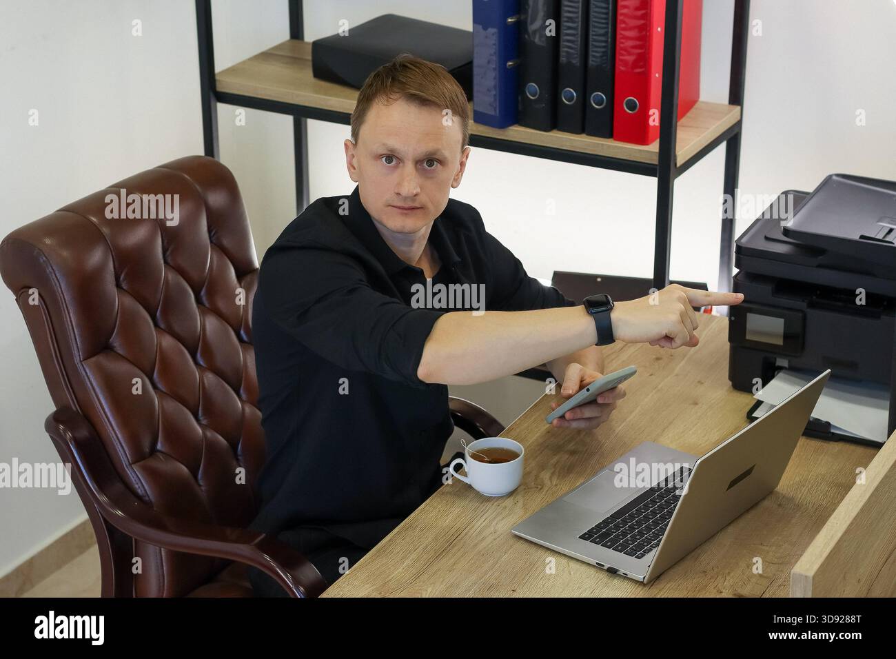 Ein professioneller Mitarbeiter sitzt am Arbeitsplatz und lenkt den Finger auf die Büroausstattung, während er mobil ist. Stockfoto