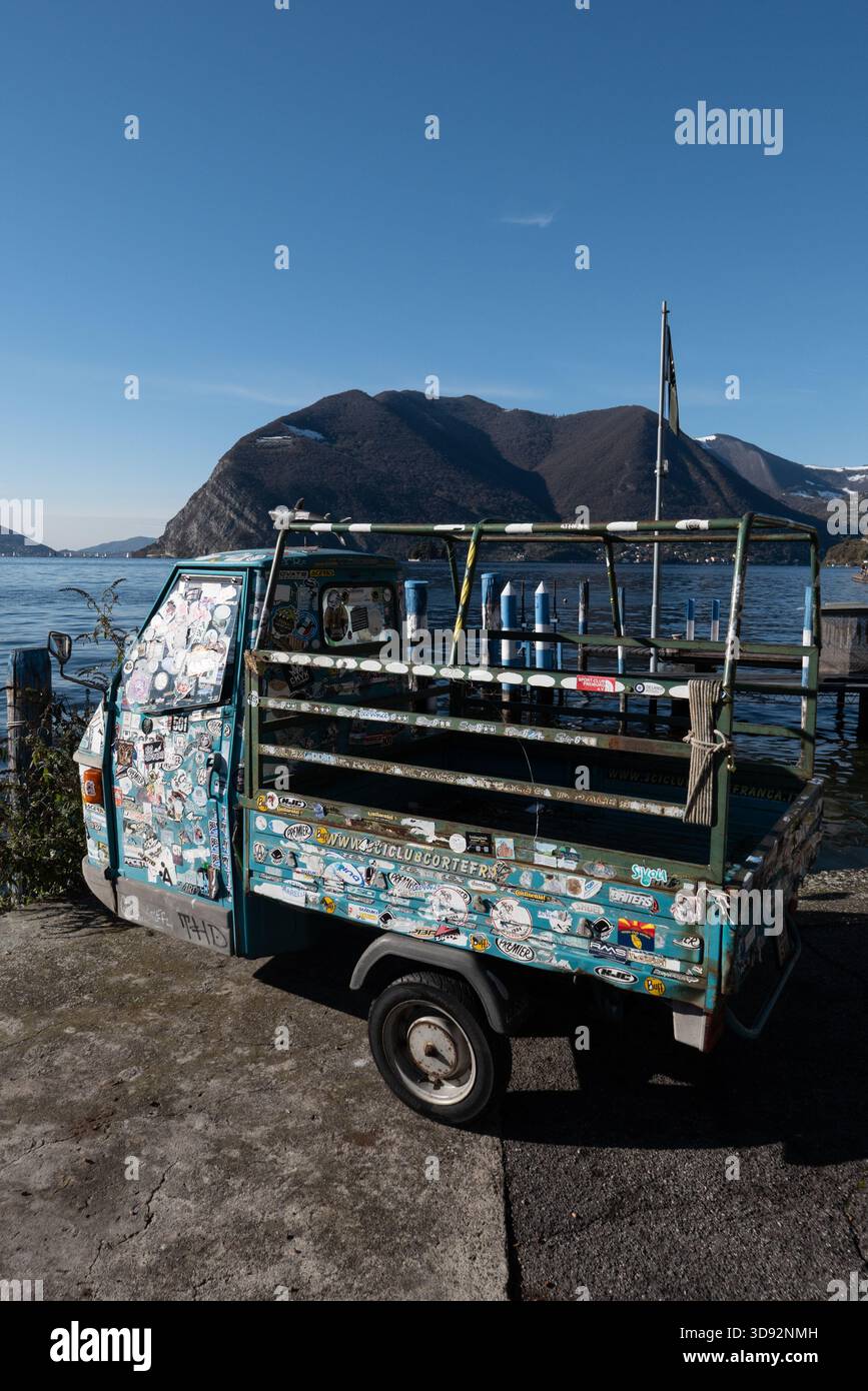 Farbenfrohe Piaggio Ape, bedeckt mit Stickern, die am Iseo-See geparkt sind, schafft eine schrullige und lebhafte Szene mit Bergen, ruhigem Wasser und einem verspielten Italiener Stockfoto