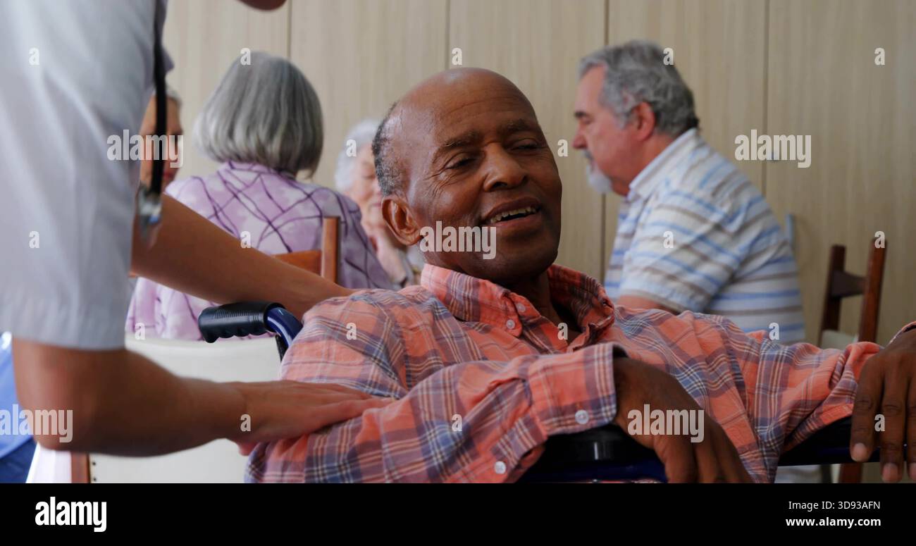 Zurückgelehnter Seniorenmann im Rollstuhl, Personal in Peelings, Hand auf Schulter im Gemeinschaftsraum Stockfoto