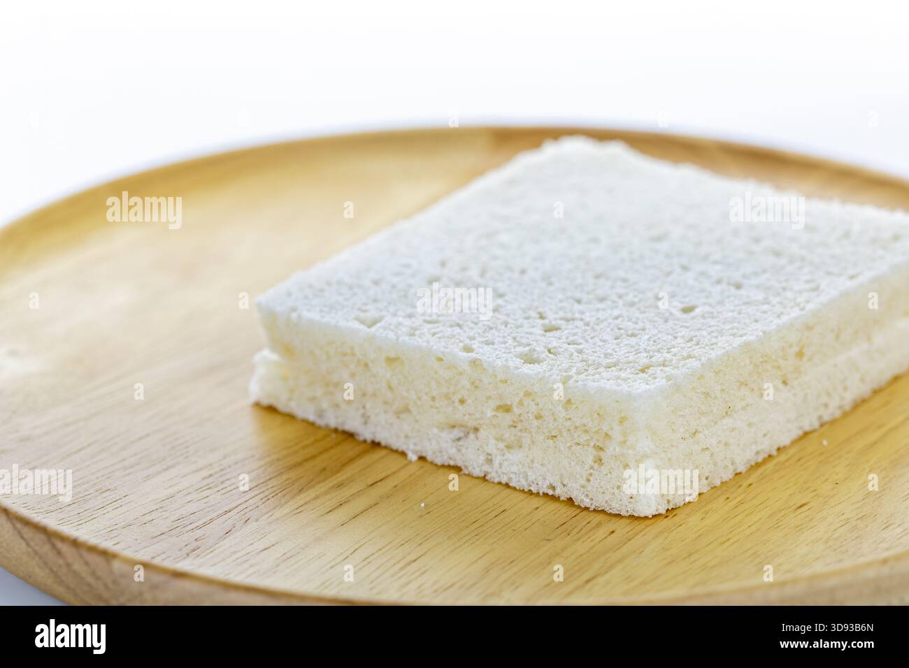 Aus nächster Nähe eine quadratische Scheibe Weißbrot auf einem Holzteller, die die weiche, poröse Textur des Brotes vor einer einfachen, verschwommenen Struktur hervorhebt Stockfoto