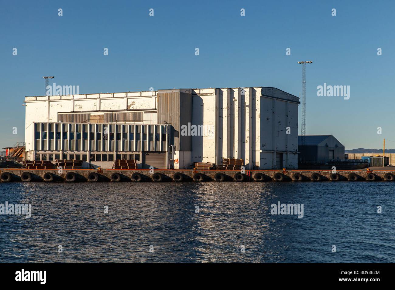 Ein großes weißes Industrielager mit Paletten und Versandausrüstung befindet sich entlang des Docks, neben ruhigem Wasser und einem klaren blauen Himmel. Maritim, logistisch Stockfoto