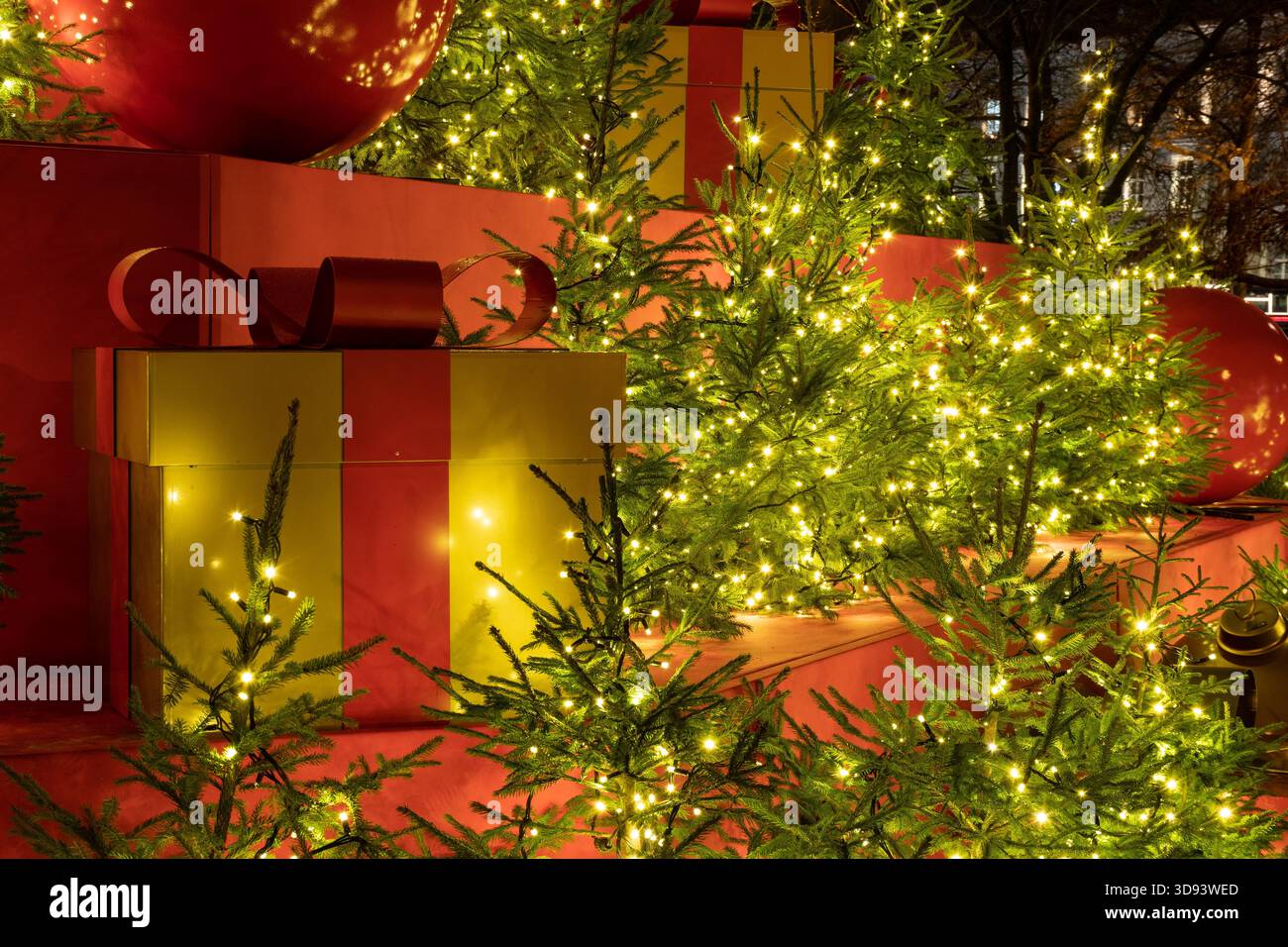 Eine große goldene Geschenkbox mit rotem Band befindet sich zwischen kleinen, verzierten Weihnachtsbäumen mit leuchtenden Lichtern Stockfoto