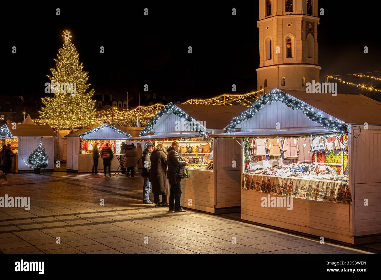 Ein hell erleuchteter Weihnachtsbaum auf einem festlichen Stadtplatz in Vilnius, Litauen, Europa, bei Nacht, umgeben von Menschen, Weihnachtsbeleuchtung und Marktstände Stockfoto