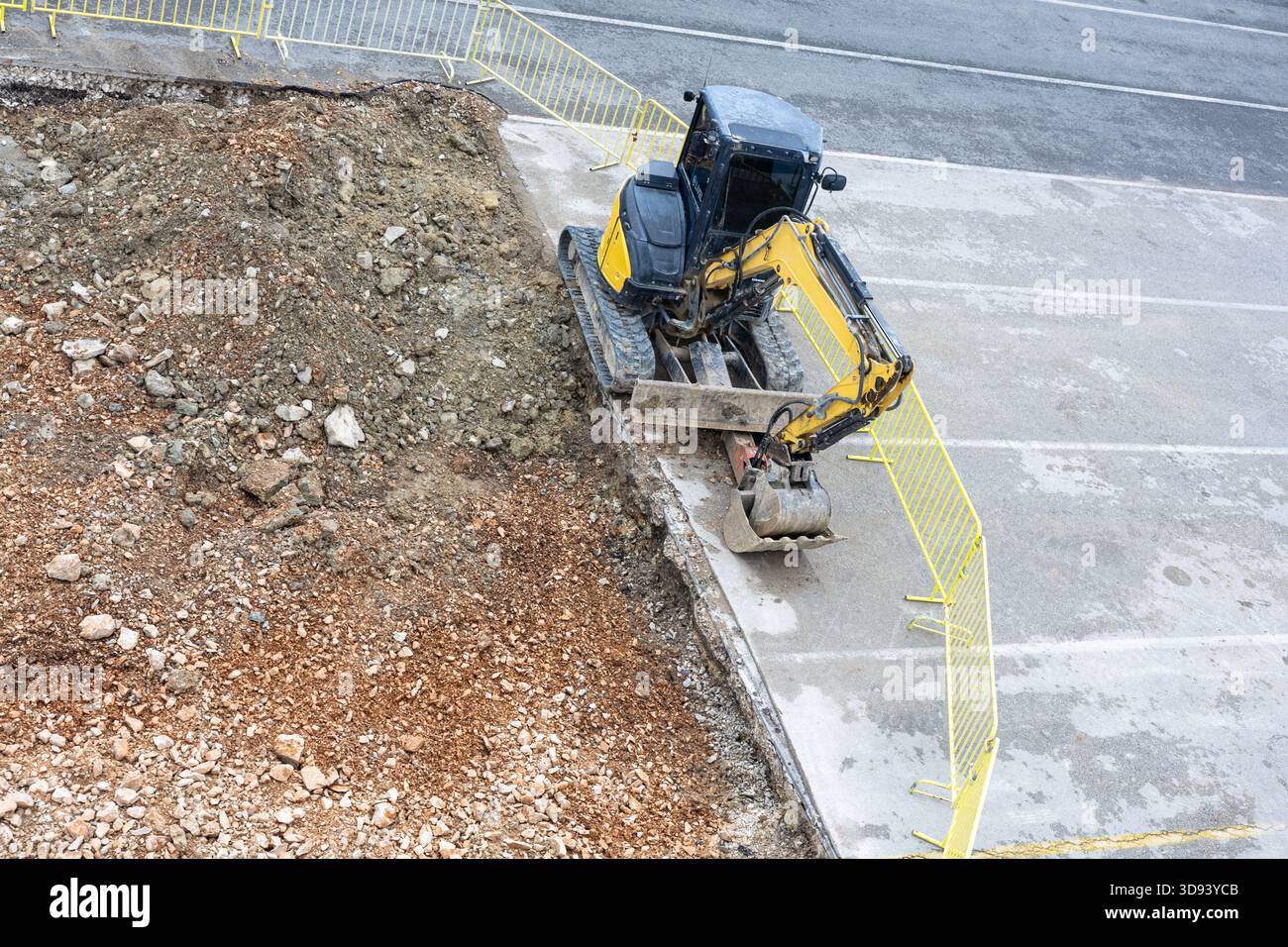 Ein Kompaktbagger gräbt einen Graben auf einer Straßenbaustelle, umgeben von hellgelben Sicherheitsbarrieren. Die Szene vermittelt urbane Entwicklung, schwer Stockfoto