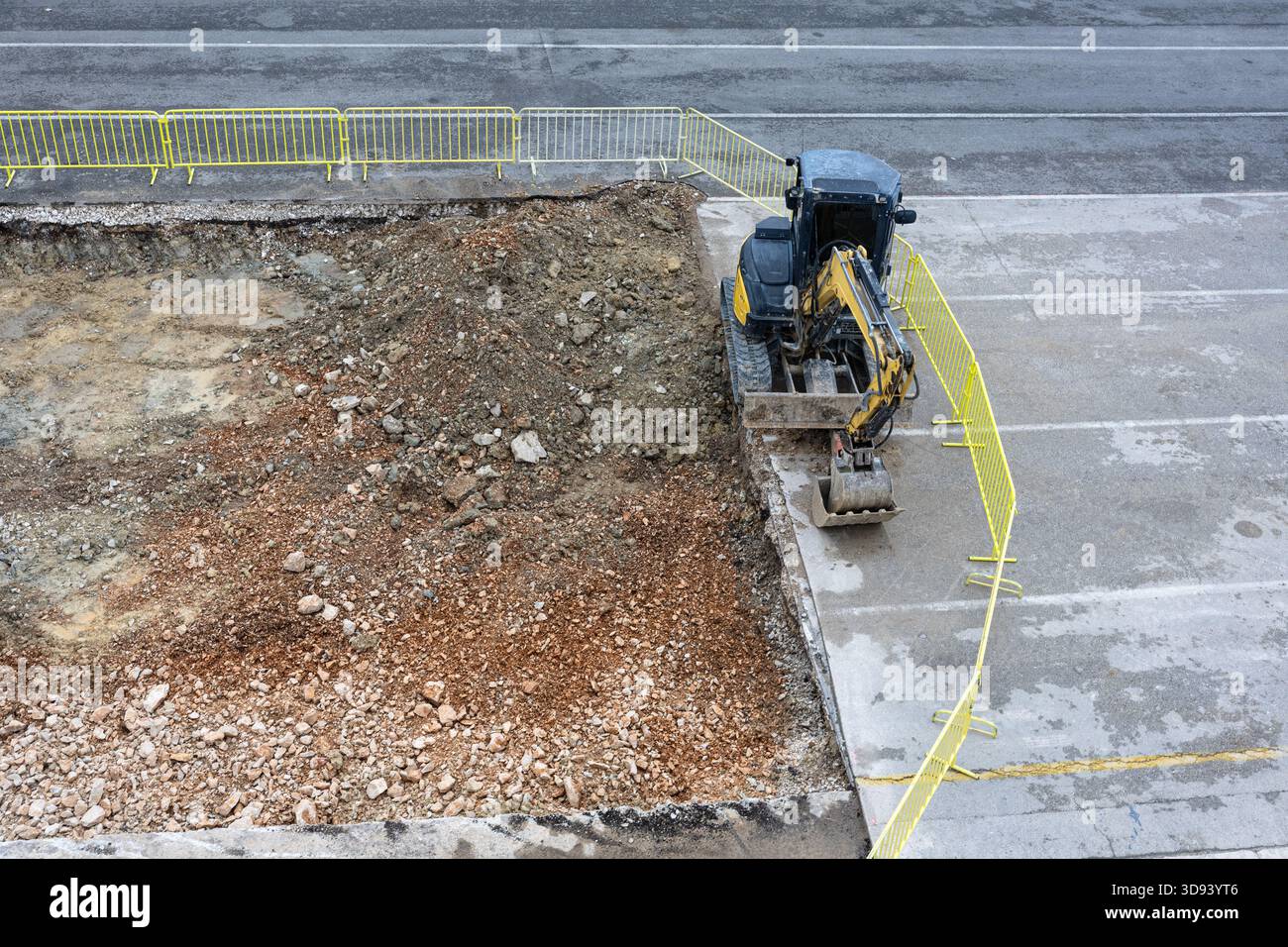 Ein Kompaktbagger gräbt einen Graben auf einer Straßenbaustelle, umgeben von hellgelben Sicherheitsbarrieren. Die Szene vermittelt urbane Entwicklung, schwer Stockfoto