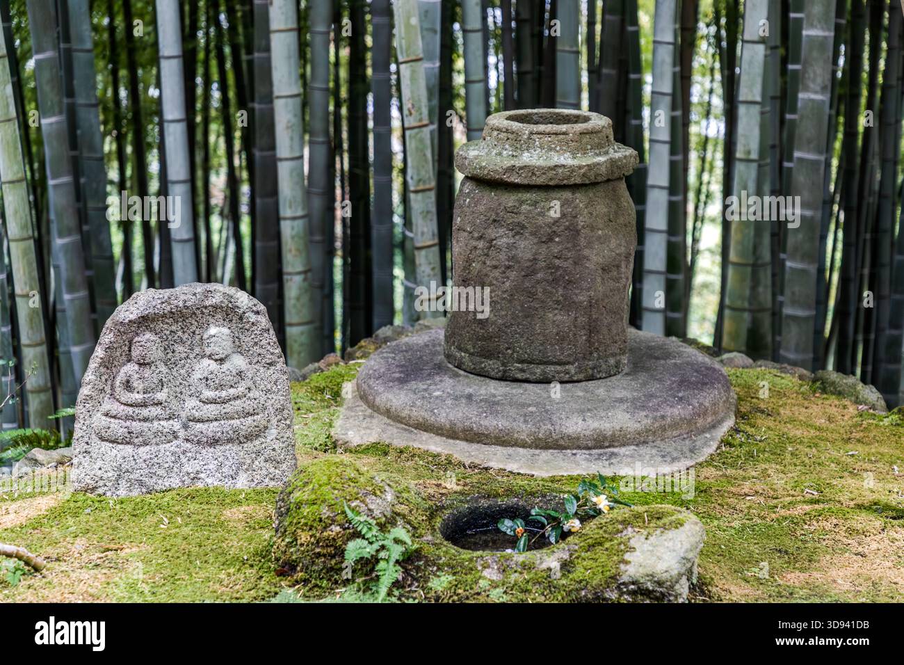 Ein buddhistisches Steinensemble in einem Bambushain in Odawara, Präfektur Kanagawa, Japan: Ein Steinrelief mit zwei sitzenden Jizo-Figuren (Bodhisattva Ksitigarbha, links), ein zylindrisches Steinobjekt auf einem runden Sockel – vermutlich ein Grab oder Reliquien – und ein Steinbecken mit Blumenopfern im Vordergrund. Der moosbedeckte Boden und die Bambusstiele im Hintergrund sind charakteristisch für diesen gut erhaltenen Tempelkomplex Stockfoto