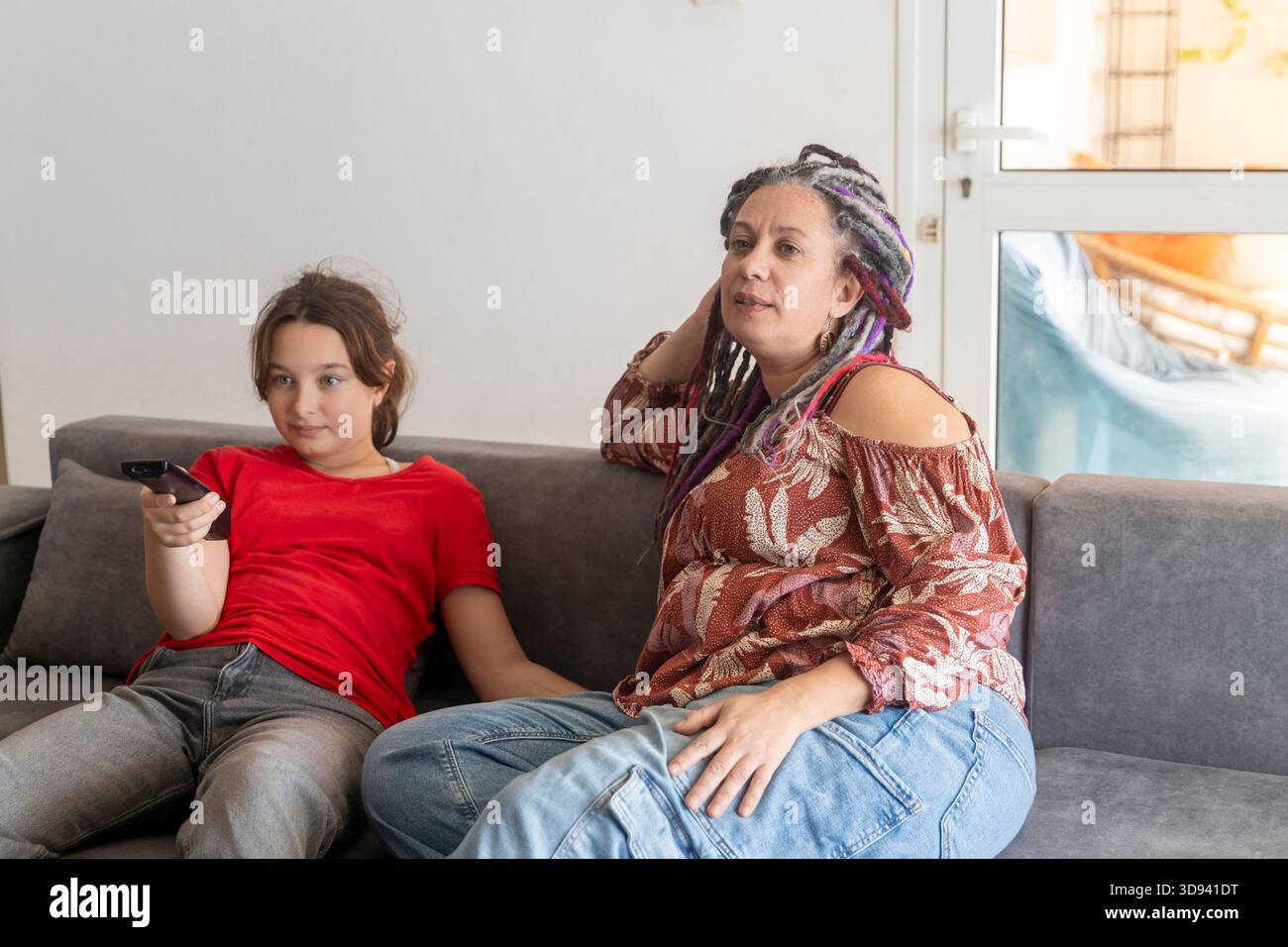 Mutter und Tochter sitzen zusammen auf einem grauen Sofa und schauen fern, während das Mädchen die Fernbedienung hält. Stockfoto