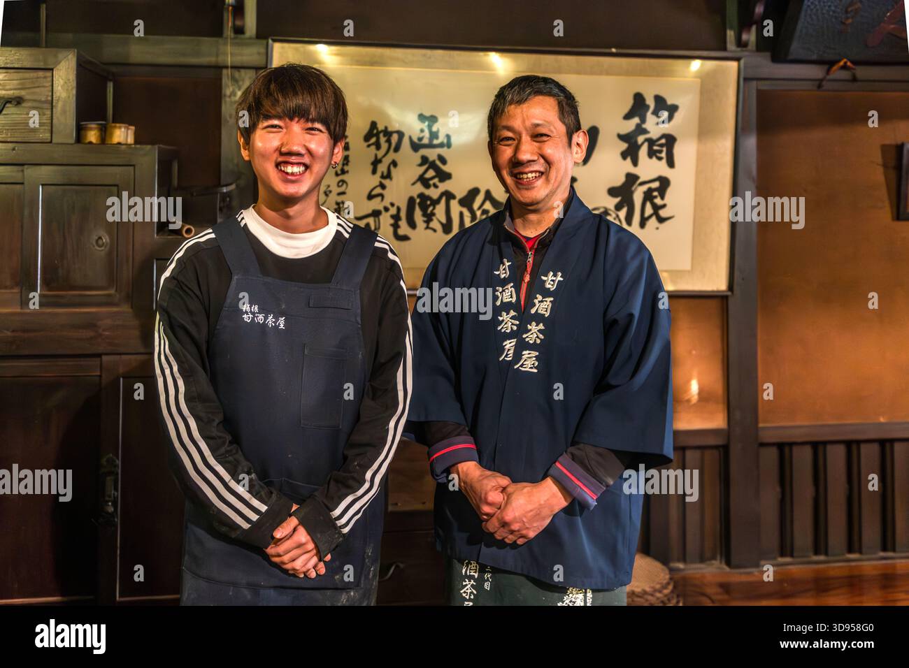 Zwei Besitzer (Vater und Sohn) des Amazake Chaya (甘酒茶屋) in Hakone, Präfektur Kanagawa, Japan, tragen traditionelle Arbeitskleidung. Im Hintergrund befindet sich eine Kalligraphie-Schriftrolle mit dem Zeichen für Hakone (箱根). Das Amazake Chaya ist eines der ältesten Teehäuser an der historischen Tokaido Road Stockfoto
