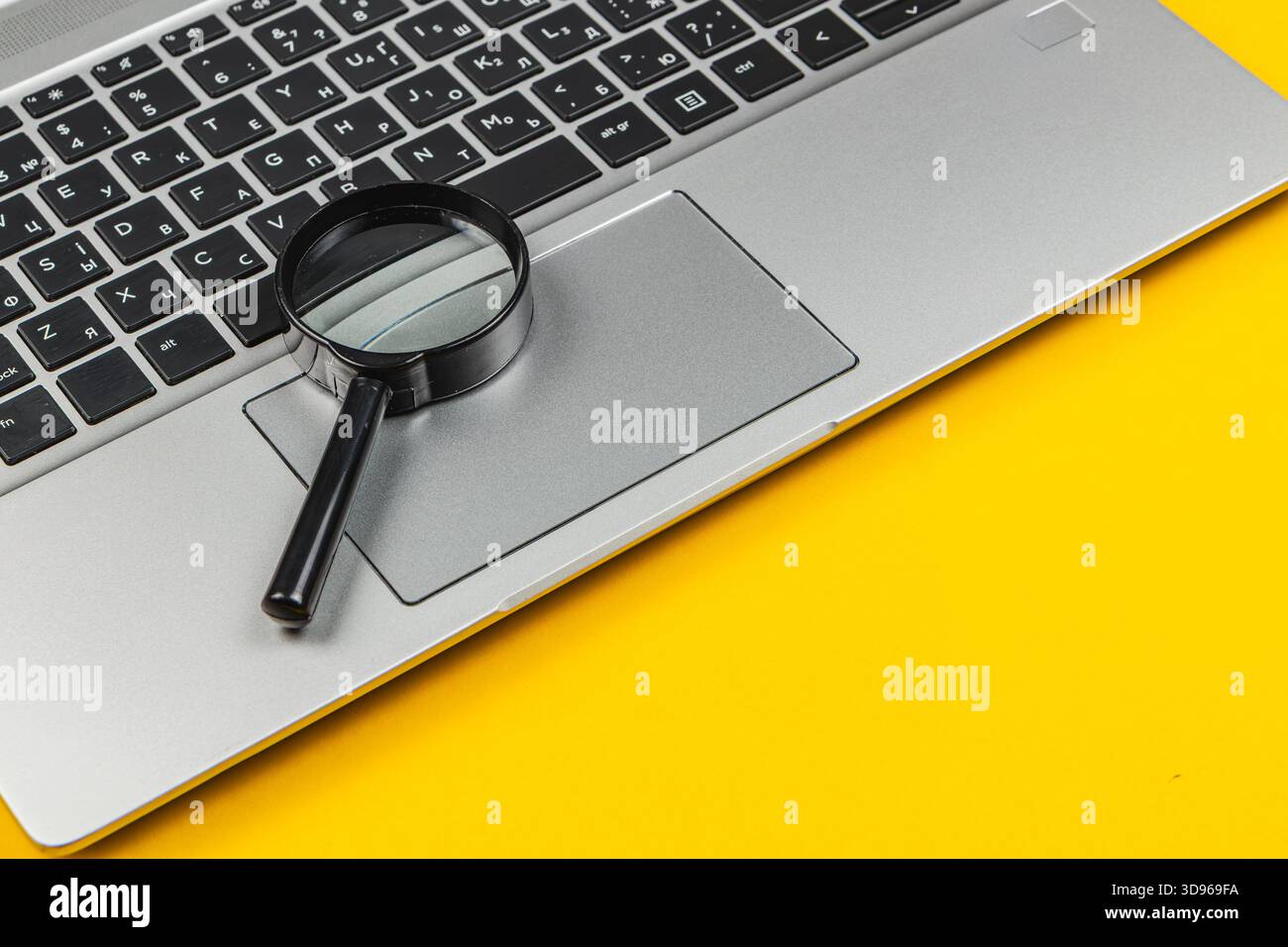 Eine Lupe liegt auf dem Touchpad eines silbernen Laptops mit leuchtendem gelbem Hintergrund, was auf Recherchen oder detaillierte Untersuchungen von Online-Ma hindeutet Stockfoto