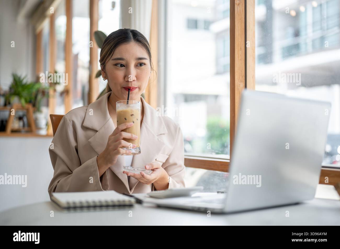 Hübsche asiatische Frau, die Glas coster hält, als Eiskaffee mit Strohhalm trinkt, während sie auf den Laptop auf dem Tisch des Cafés blickt. Arbeiten oder Lernen, Kühlen im Freien Stockfoto