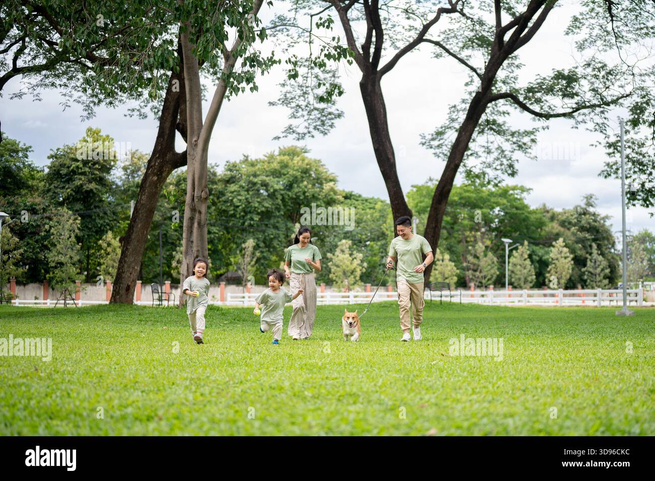 Dad hält die Leine an einem Corgi-Welpen, während er mit Mutter und Tochter auf Gras im Park läuft. Eltern und Kinder, Hund, Familienausflug. Stockfoto