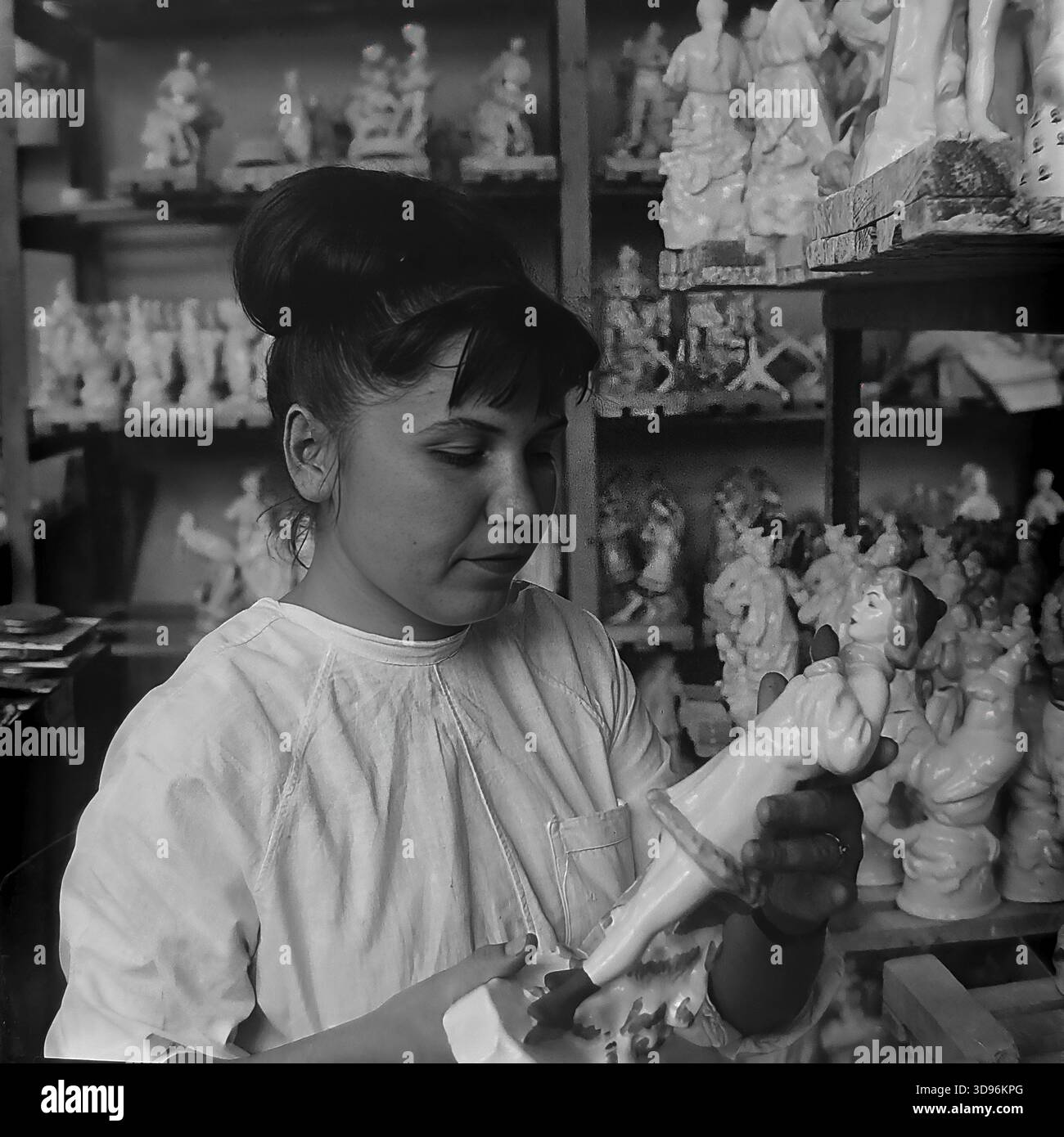Slawjansk, Ukraine, UdSSR. 1968. Schwarzweiß-Archivfoto, auf dem eine Angestellte des Keramikkomplexes Slavjansk die Qualitätskontrolle von Porzellanerzeugnissen durchführt. Die Frau im weißen Arbeitsmantel inspiziert eine kleine Figur in ihren Händen. Hinter ihr befinden sich Holzregale mit Reihen identischer Statuetten, die Mädchen in Wintermänteln und -Muffen darstellen. Das Bild zeigt die Massenproduktion von Souvenirs und die Liebe zum Detail im friedlichen Donbass vor dem Krieg. Stockfoto