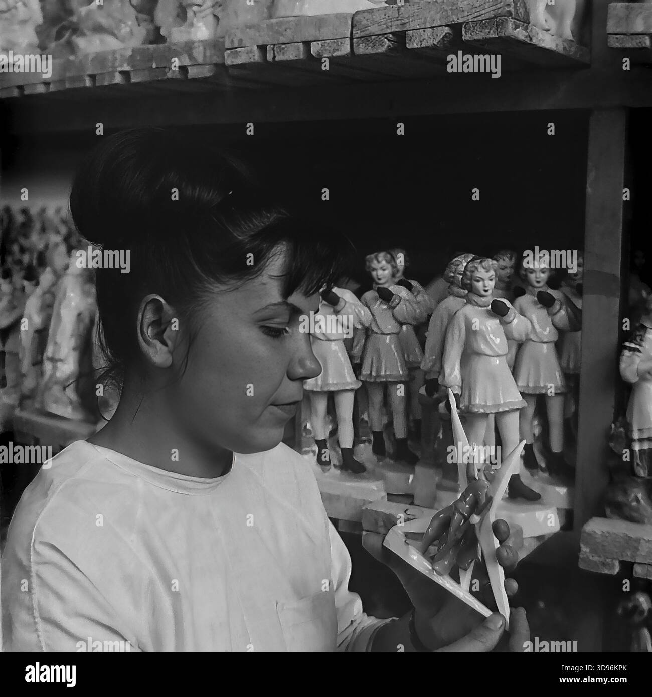 Slawjansk, Ukraine, UdSSR. 1968. Schwarzweiß-Archivfoto, auf dem eine Angestellte des Keramikkomplexes Slavjansk die Qualitätskontrolle von Porzellanerzeugnissen durchführt. Die Frau im weißen Arbeitsmantel inspiziert eine kleine Figur in ihren Händen. Hinter ihr befinden sich Holzregale mit Reihen identischer Statuetten, die Mädchen in Wintermänteln und -Muffen darstellen. Das Bild zeigt die Massenproduktion von Souvenirs und die Liebe zum Detail im friedlichen Donbass vor dem Krieg. Stockfoto