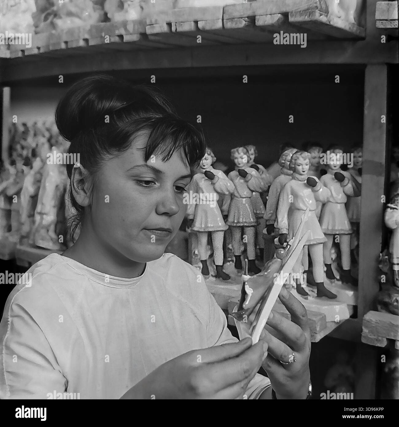 Slawjansk, Ukraine, UdSSR. 1968. Schwarzweiß-Archivfoto, auf dem eine Angestellte des Keramikkomplexes Slavjansk die Qualitätskontrolle von Porzellanerzeugnissen durchführt. Die Frau im weißen Arbeitsmantel inspiziert eine kleine Figur in ihren Händen. Hinter ihr befinden sich Holzregale mit Reihen identischer Statuetten, die Mädchen in Wintermänteln und -Muffen darstellen. Das Bild zeigt die Massenproduktion von Souvenirs und die Liebe zum Detail im friedlichen Donbass vor dem Krieg. Stockfoto