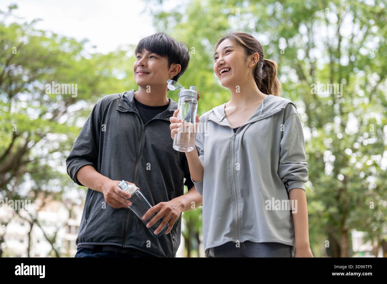 Asiatische Männer und Frauen in Sportbekleidung, die eine Wasserflasche halten und in die gleiche Richtung auf den Innenhof des Parks schauen. Junges Paar, Trainingsanzug. Grüne Natur. Stockfoto