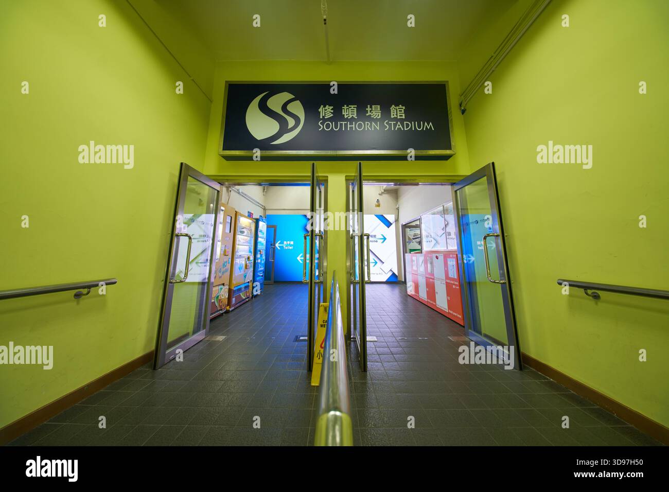 Der Eingang am 1/F zum Southorn Stadium mit hellgrünen Wänden, Glastüren und Beschilderung in chinesischer und englischer Sprache. Stockfoto