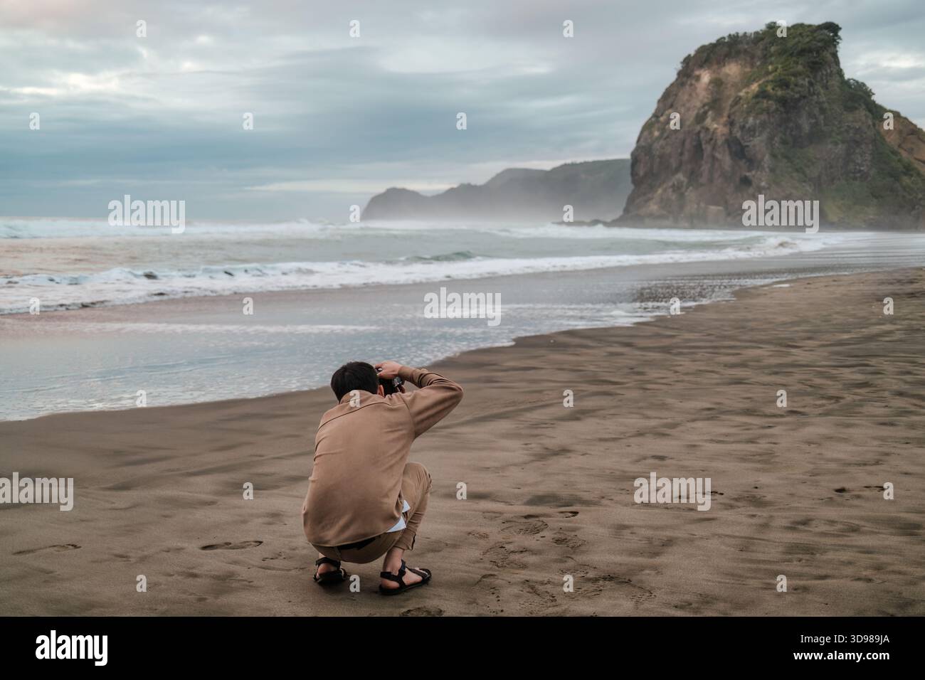 Männlicher Tourist, der ein Foto mit Lion Rock und Meereswelle am Piha Beach in der Westküste von Auckland, Neuseeland macht Stockfoto
