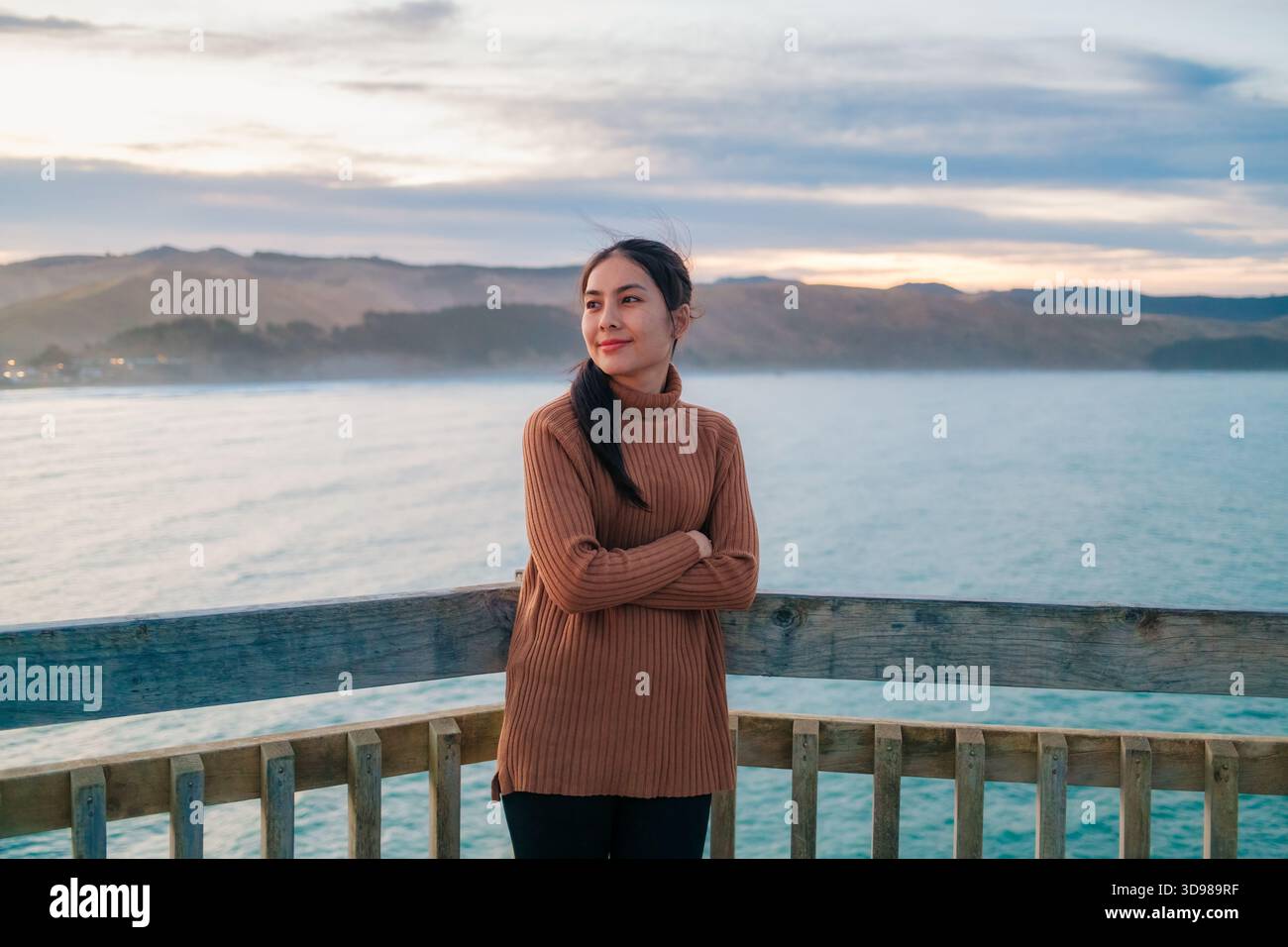 Schöne asiatische Frau in braunem Pullover am pazifik mit Bergblick bei Sonnenuntergang in Wellington, Neuseeland Stockfoto