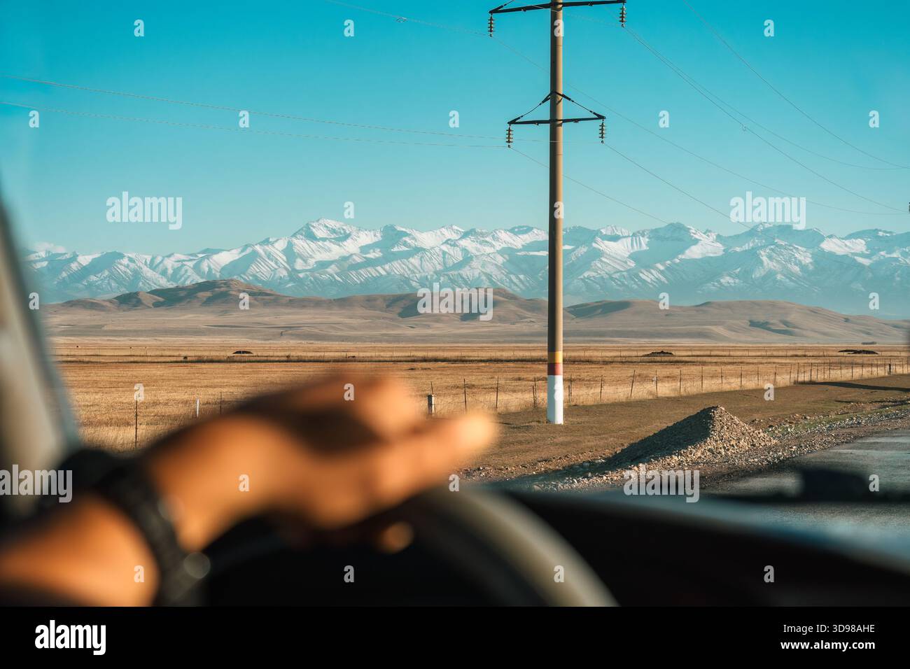Roadtrip mit Fahrer, der das Lenkrad hält, während er die offene Ebene mit Stromleitungen und schneebedeckten Bergen unter blauem Himmel in der Nähe der Grenze von Ky überquert Stockfoto