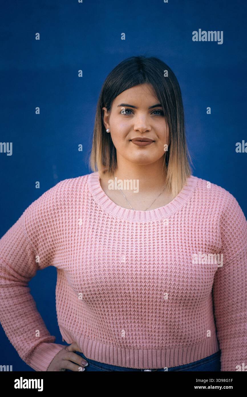 Porträt einer jungen Frau mit glatten Haaren, die einen rosa Pullover trägt und selbstbewusst vor einer blauen Wand steht. Stockfoto