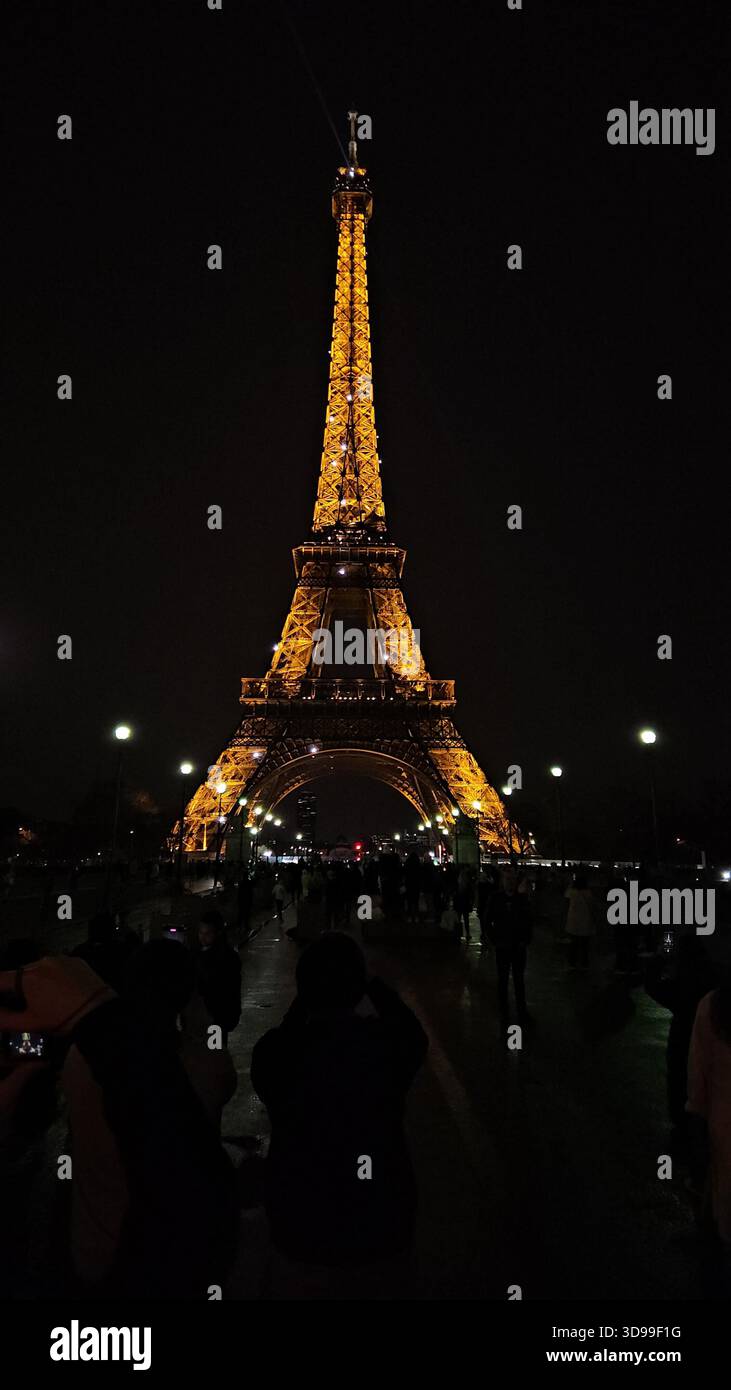 Der Eiffelturm leuchtet nachts in Paris, Frankreich, mit Reflexionen auf nassem Gehweg und lebhafter Menschenmenge. Stockfoto