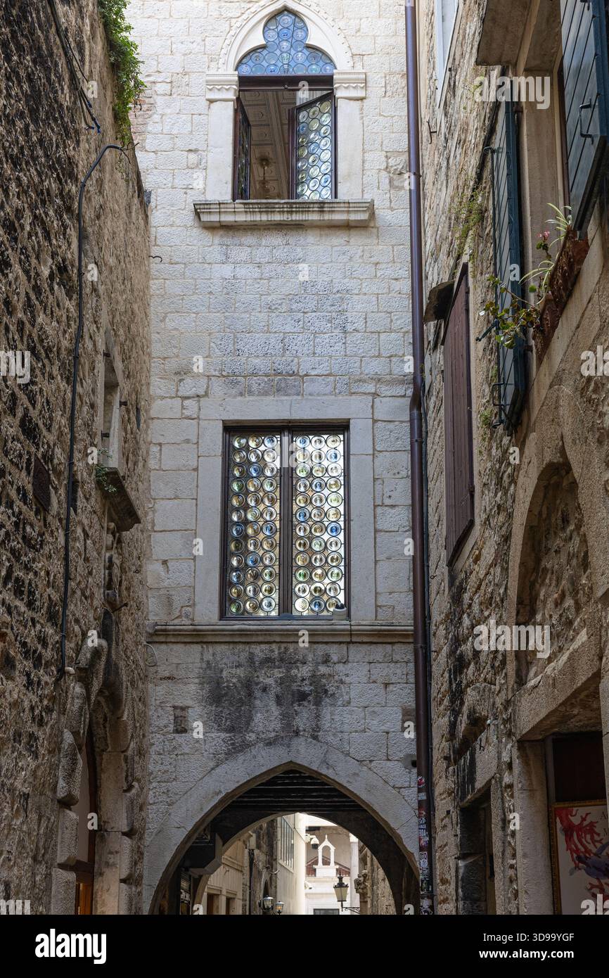Eine ruhige, historische Gasse, umgeben von verwitterten Steinmauern. Ein bogenförmiges Fenster mit runden Glasscheiben überblickt einen sonnendurchfluteten Gang und erinnert an mittelalterlichen Charme Stockfoto