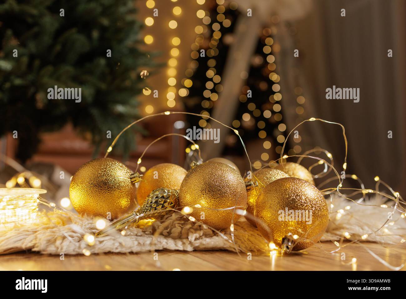 Goldene Kugeln und leuchtende Lichterketten liegen auf weichem Teppich unter Weihnachtsbaum mit warmem Bokeh in gemütlicher festlicher Inneneinrichtung. Stockfoto
