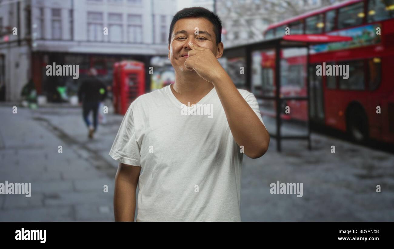 Hispanischer junger Mann im weißen T-Shirt, der seine Nase mit dem Zeigefinger berührt, auf einer Stadtstraße neben rotem Doppeldeckerbus und Telefonbox; Verspieltheit. Stockfoto
