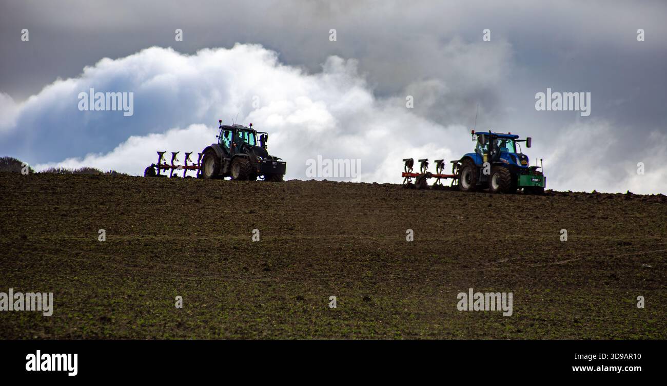 Zwei Traktoren pflügen ein großes Feld unter einem dramatisch bewölkten Himmel während der landwirtschaftlichen Feldarbeit in der frühen landwirtschaftlichen Saison Stockfoto