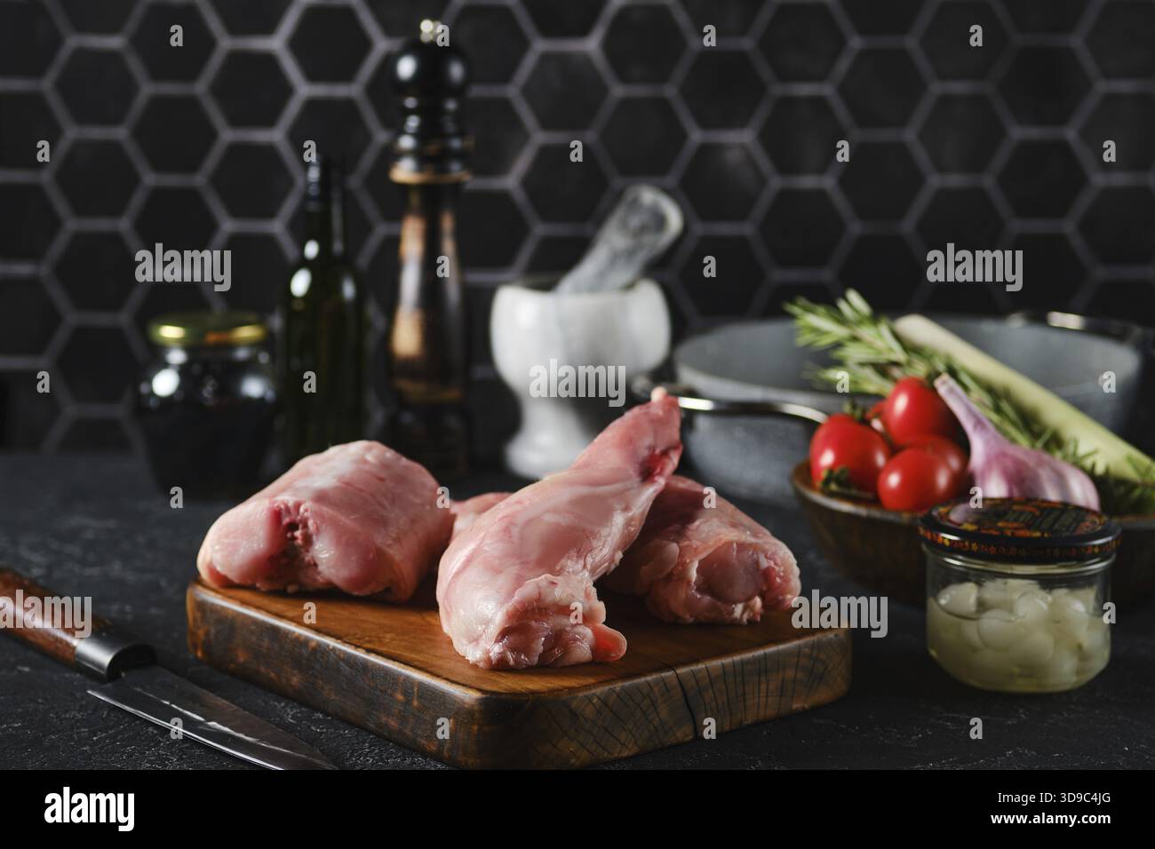 Zwei rohe Hasenbeine und Sattel auf einem hölzernen Schneidebrett, umgeben von Kochzutaten wie Kirschtomaten, Kräutern und Gewürzen Stockfoto