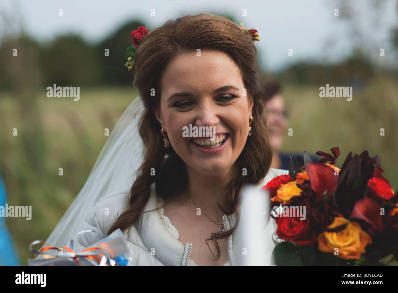 Die junge Braut lächelt hell, während sie ihren Hochzeitsstrauß im Freien in Lettland hält, mit weichem Hintergrund auf dem Land. Stockfoto