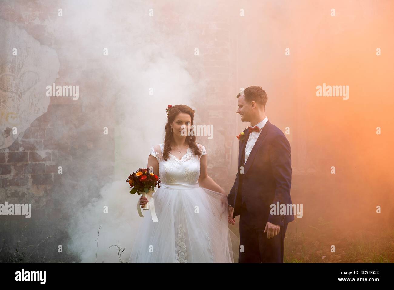 Der junge Bräutigam betrachtet die Braut, während sie in dickem, farbigem Rauch steht, und schafft ein ruhiges und emotionales Hochzeitsporträt mit Wärme, Tiefe und ruhiger Verbindung Stockfoto
