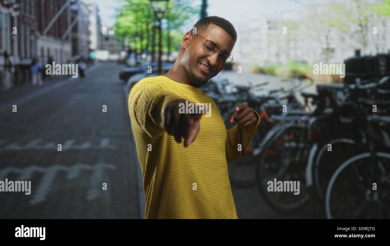 Junger schwarzer Mann, der mit dem Finger auf die Kamera zeigt und eine Fingerpistolen-Geste macht, lächelt in einem gelben Pullover auf einer Straße, die von geparkten Fahrrädern und Fahrrädern gesäumt ist Stockfoto