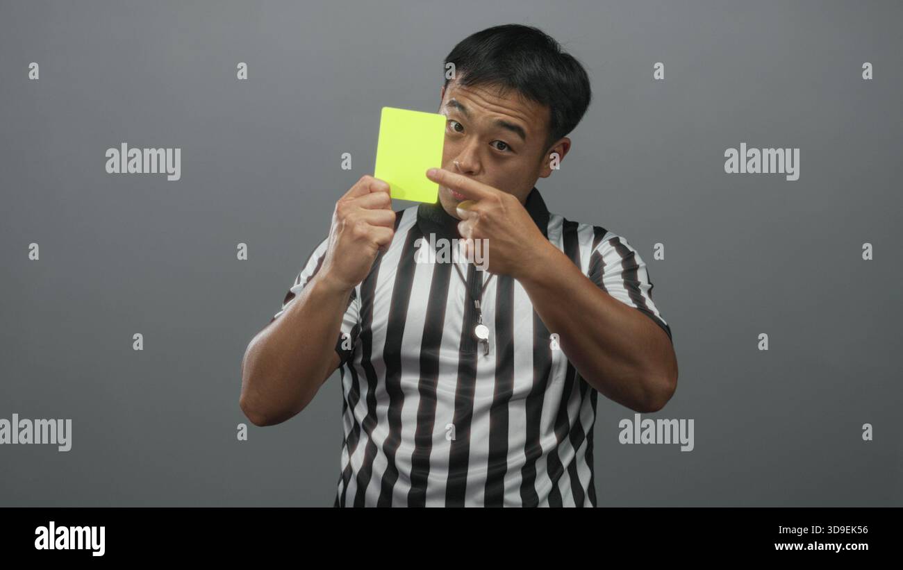 Junger chinesischer Schiedsrichter hält gelbe Karte und zeigt mit dem Finger, während er ein gestreiftes Hemd im grauen Studio trägt; Autorität. Stockfoto